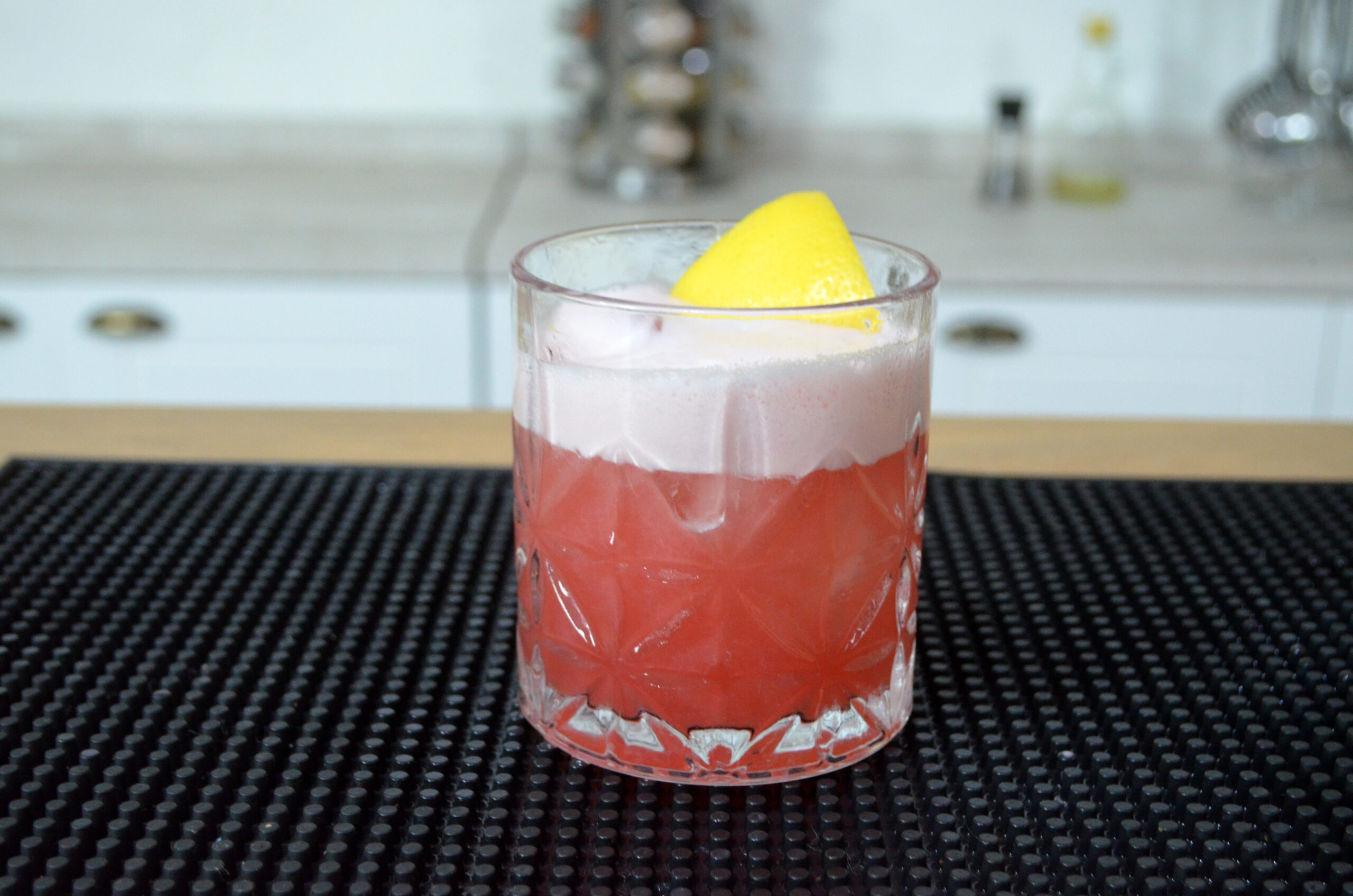 Copo de Campari Sour com espuma no topo, decorado com uma casca de limão siciliano, sobre uma mesa com ingredientes frescos ao redor.