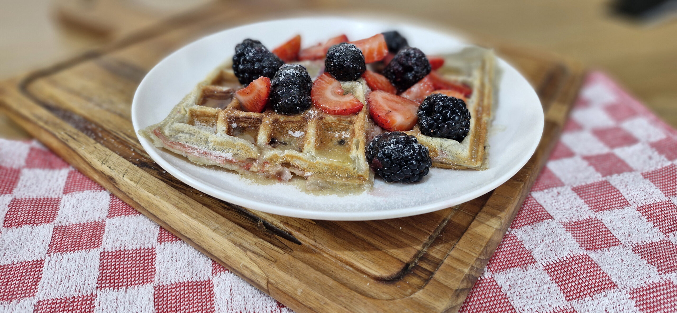 Prato com waffles dourados, regados com mel e decorados com frutas frescas como morangos e mirtilos, acompanhado de uma xícara de café ao fundo.