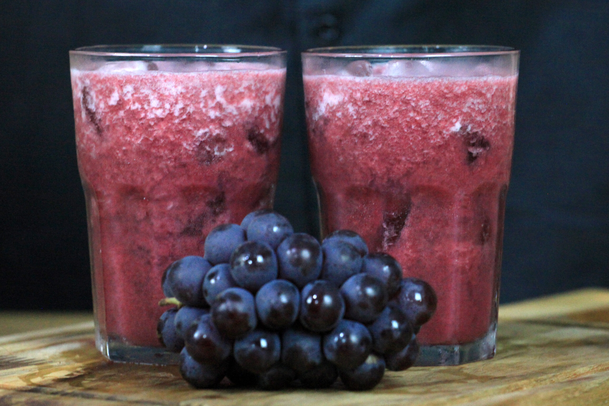 Copo de suco de uva integral, sem açúcar, com uma jarra ao fundo e uvas frescas decorando a mesa.