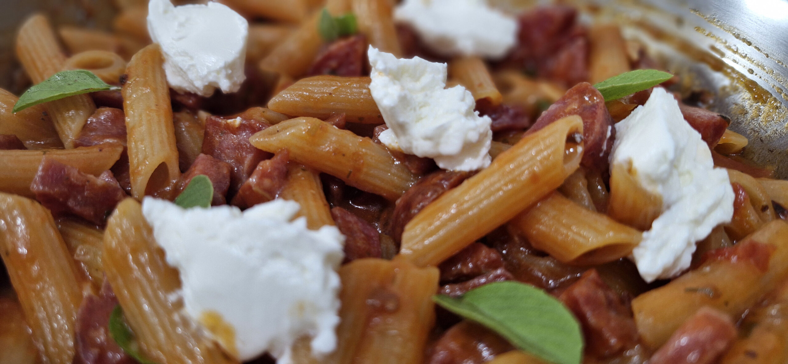 Prato de macarrão al dente com linguiça Blumenau dourada, envolvido em um molho saboroso, finalizado com ervas frescas e queijo ralado.